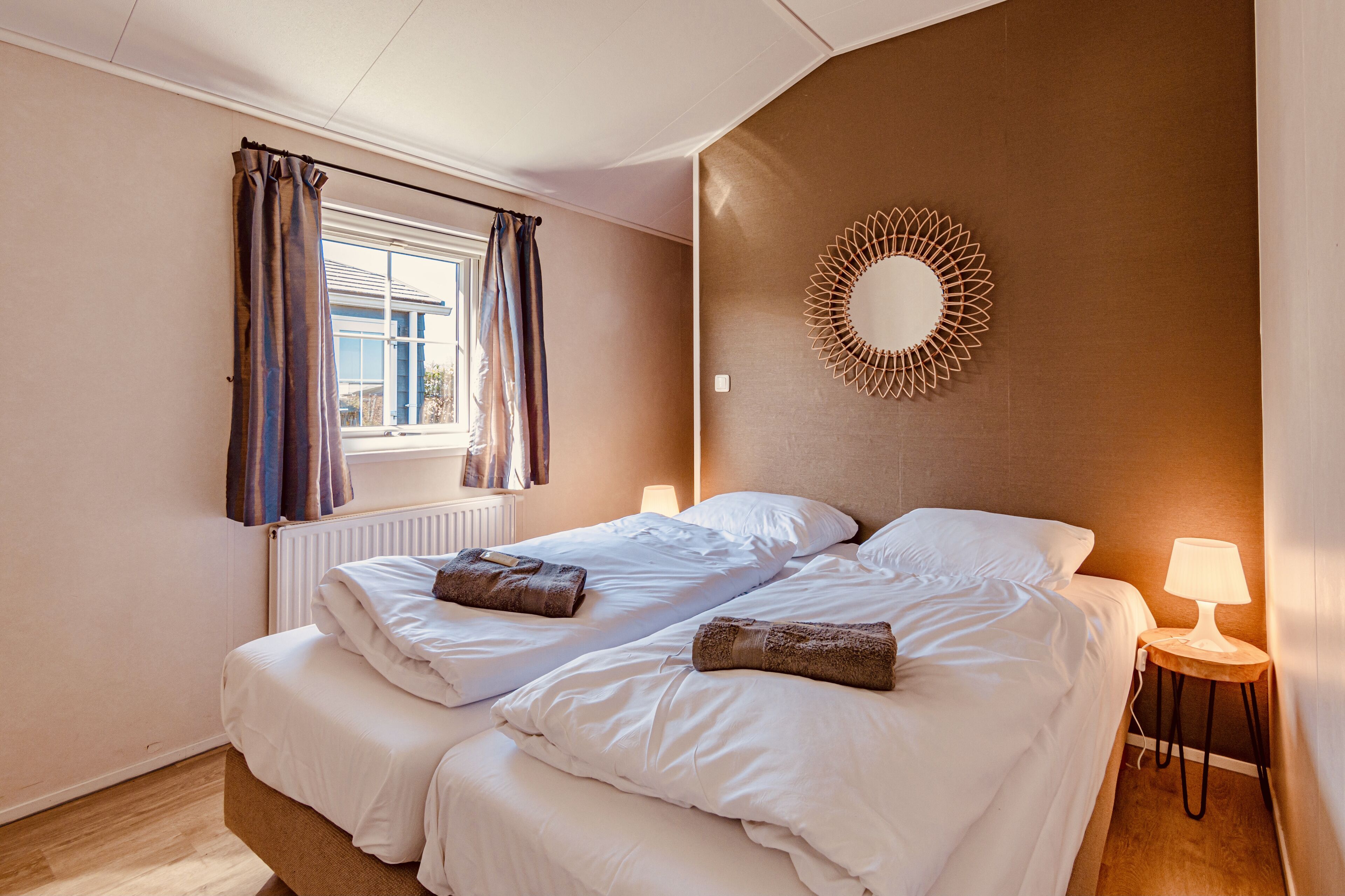A hotel room with two beds, a window with curtains, a wall-mounted mirror, and a bedside lamp.