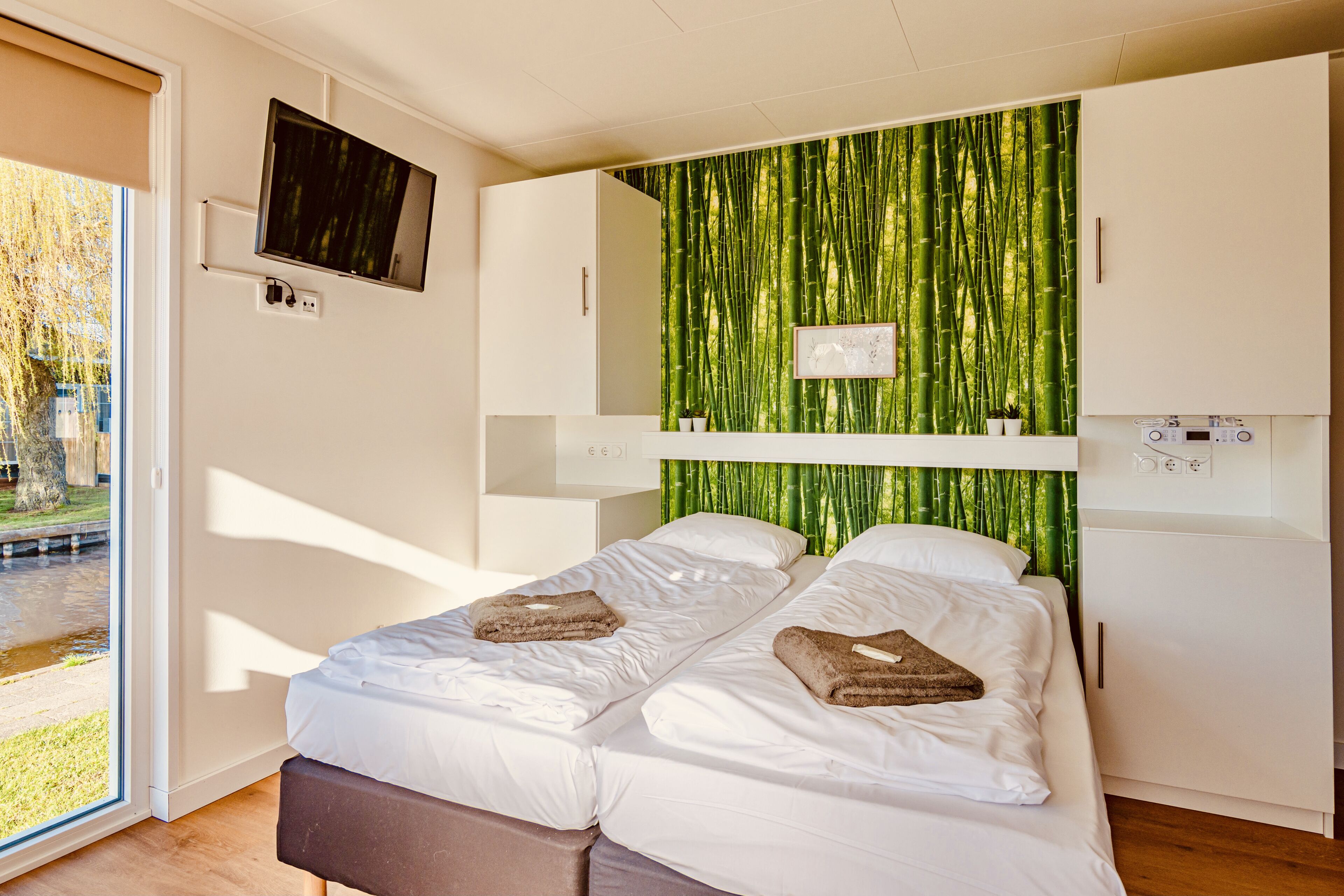 A modern hotel room with a bed, a television, a bamboo wall, and a small kitchenette.