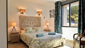 Een slaapkamer met een groot bed, twee bedlampjes, een decoratief hoofdeinde, een raam met gordijnen en een stoel met een boek.