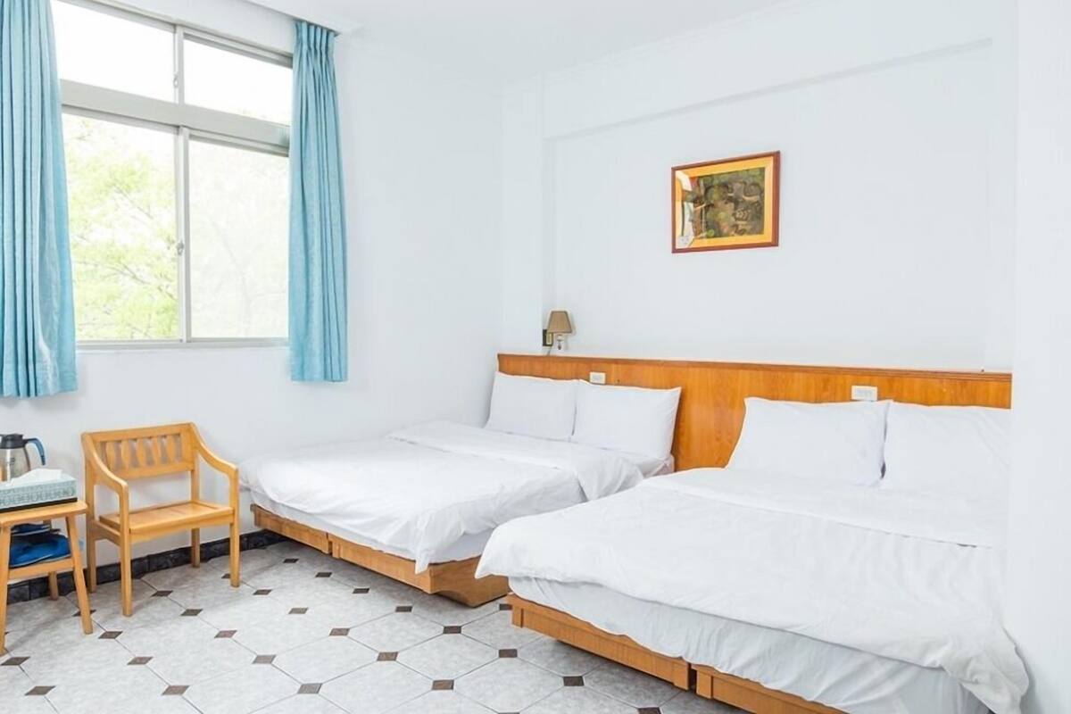 Quarto de hotel com duas camas de solteiro, uma escrivaninha de madeira, uma cadeira e uma janela com cortinas.