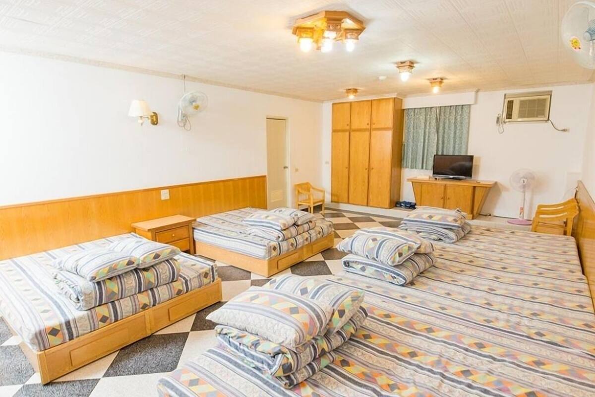 Quarto de hotel com quatro camas, televisão, guarda-roupa e secretária com cadeira.