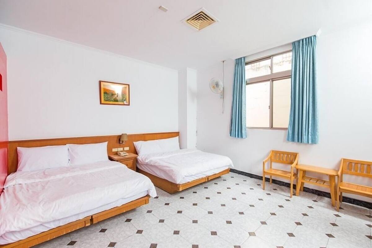 Quarto de hotel com duas camas de solteiro, uma mesinha, duas cadeiras, uma janela com cortinas e um quadro na parede.