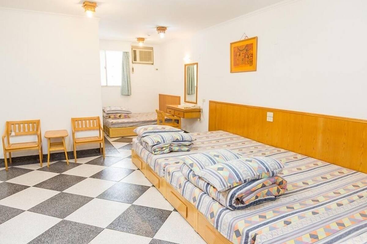 Quarto com piso xadrez, cama com roupa de cama listrada, cabeceira de madeira, mesa de cabeceira e uma cadeira.