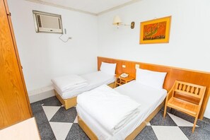 Habitación de hotel con dos camas, cabecero de madera, mesita de noche con lámpara, una silla, un cuadro en la pared y una ventana con aire acondicionado.