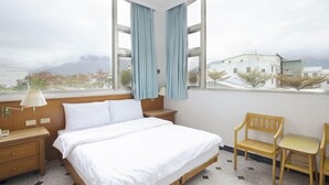 Habitación de hotel con cama, mesita de noche, lámpara, silla y vistas a edificios y montañas a través de amplias ventanas.