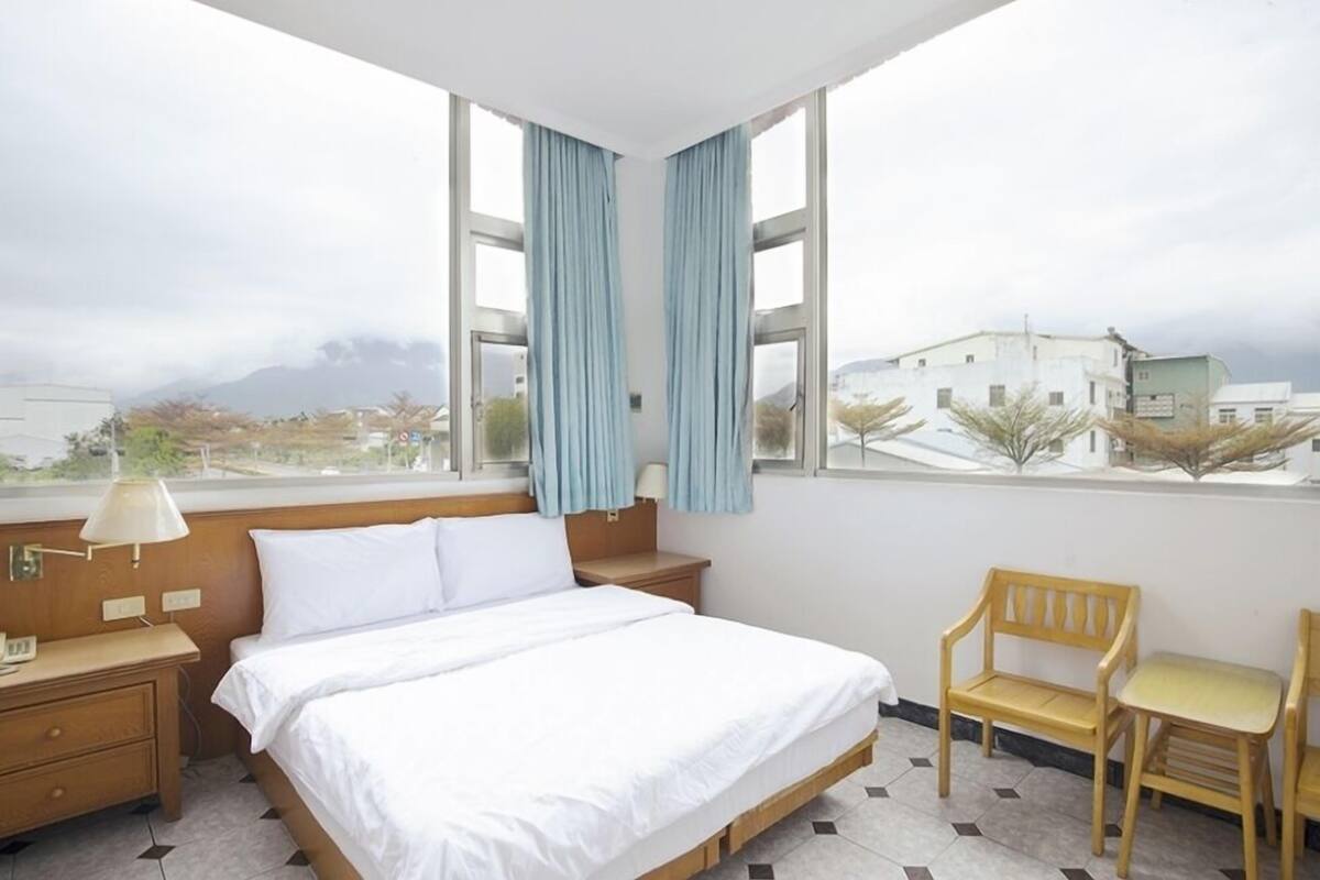Quarto de hotel com cama, mesa de cabeceira, luminária, cadeira e vista para edifícios e montanhas através de grandes janelas.