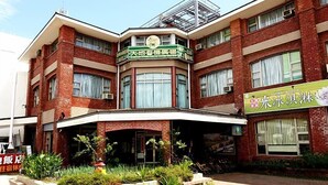 Un edificio de varios pisos con fachada de ladrillo, un letrero verde y una zona de acceso cubierta con plantas.