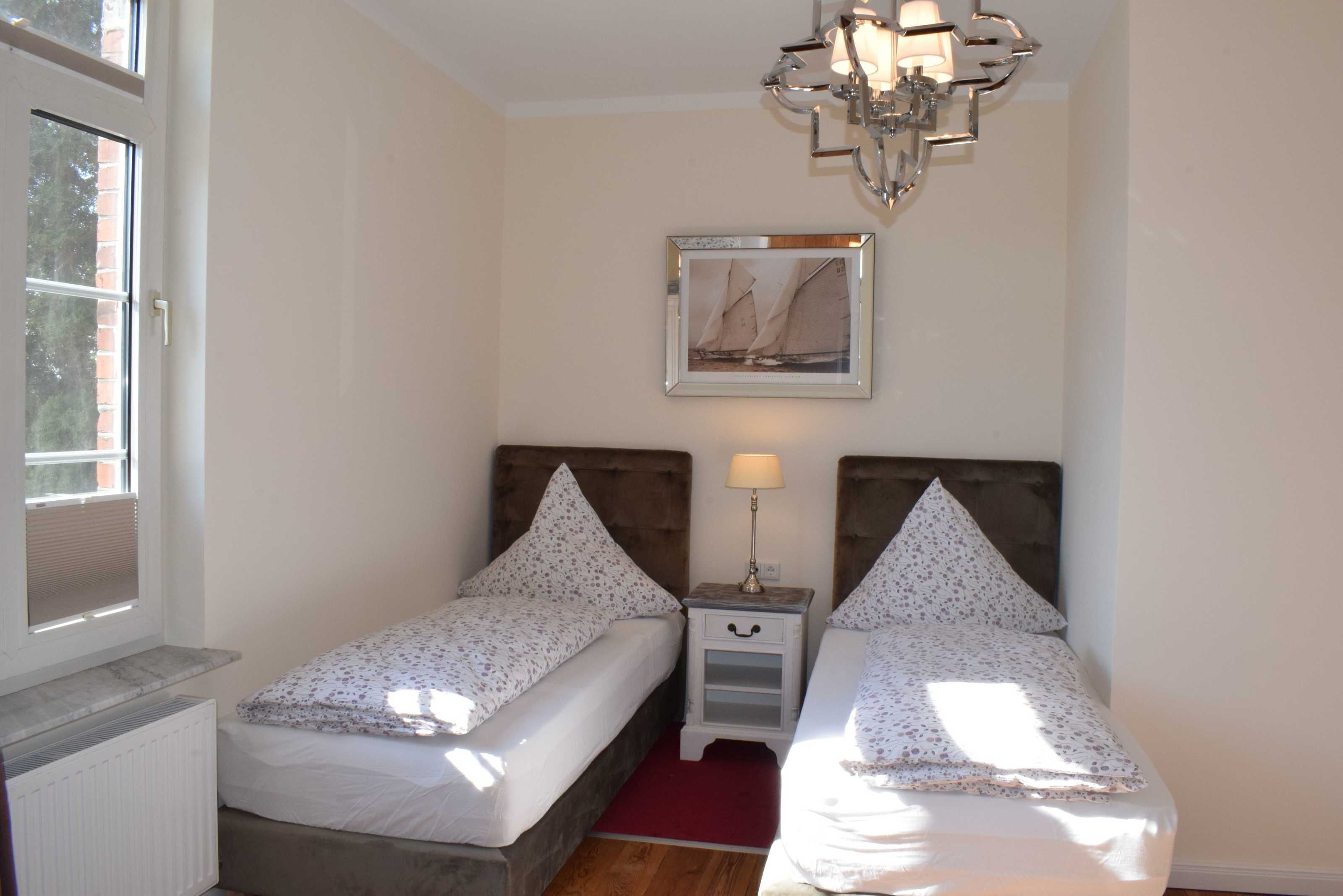 A bedroom with two single beds, a nightstand with a lamp, a window with blinds, and a framed picture on the wall.