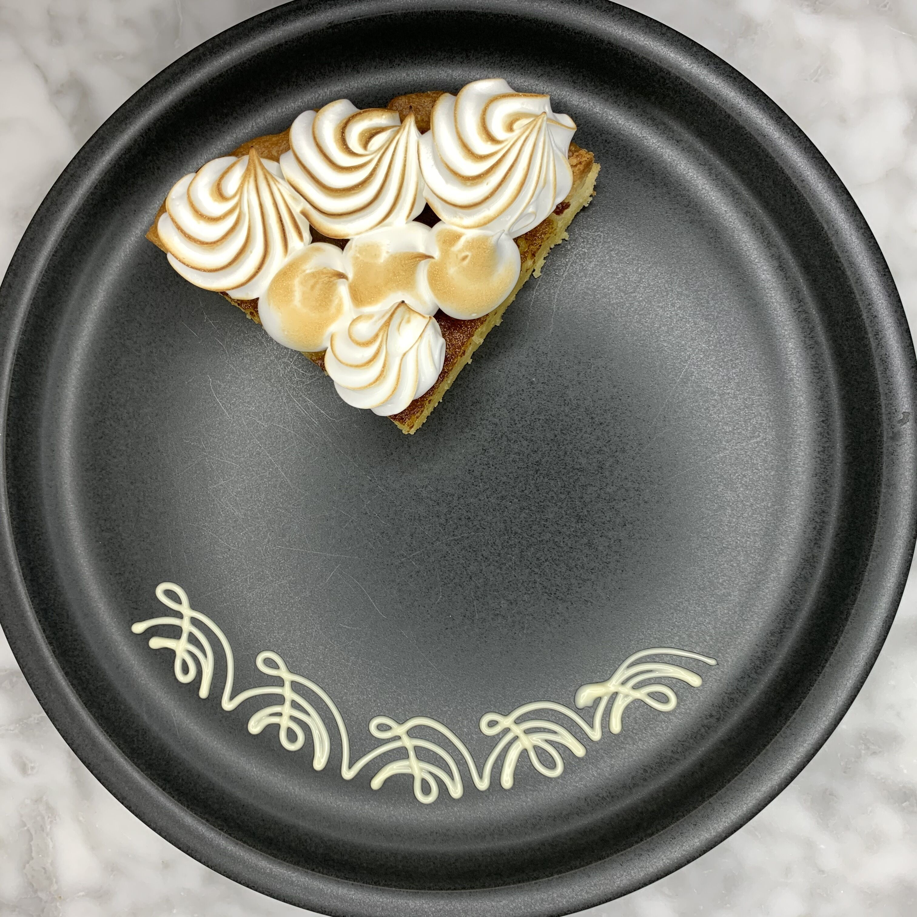 Una porción de pastel con crema batida sobre un plato negro con motivos de hojas decorativas.
