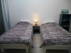 A hotel room with two beds, each with a grey bedspread featuring the text "New York City". There is a nightstand with a lamp between the beds, and a bookshelf to the right. - Apartment with terrace, parking and port view in Capbreton (Capbreton)