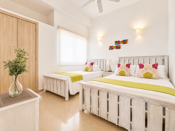 Une chambre d’hôtel avec deux lits, une fenêtre avec des stores, un radiateur, un vase avec des fleurs et un luminaire fixé au mur.