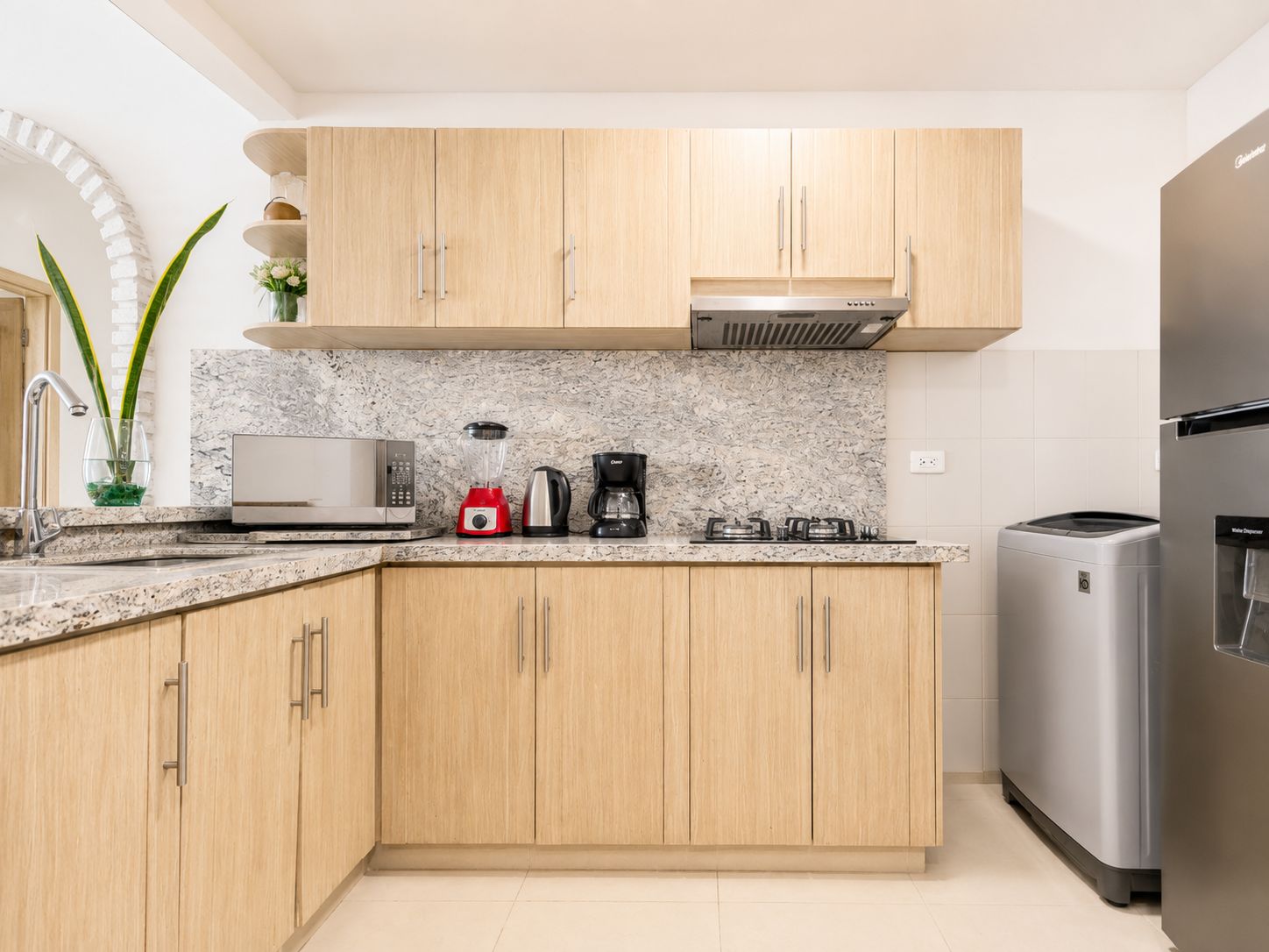 A modern kitchen with wooden cabinets, a granite countertop, a built-in microwave, a red blender, a coffee maker, and a stainless steel refrigerator.