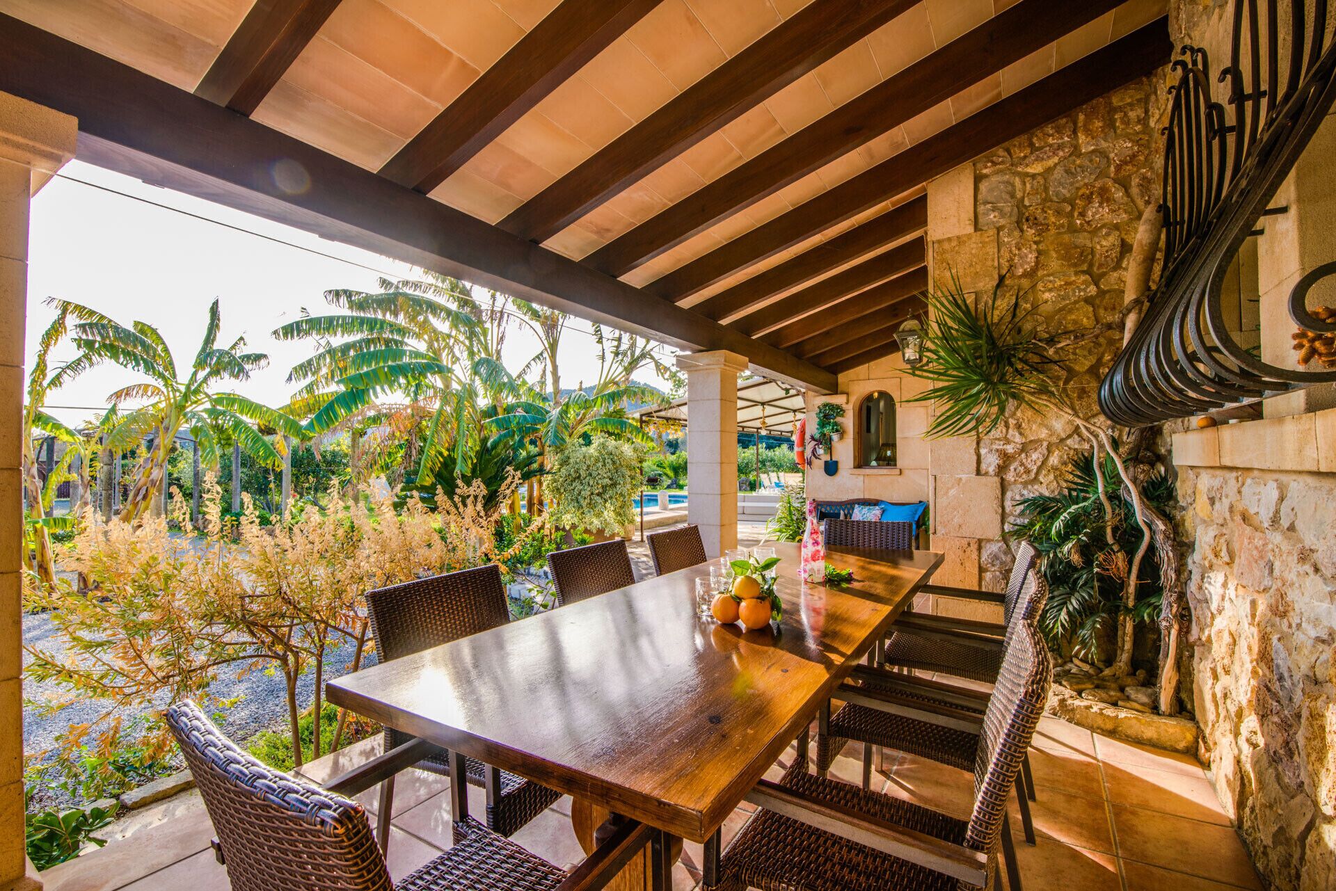 Un patio al aire libre con una mesa de madera y sillas de mimbre, rodeado de exuberante vegetación tropical y un muro de piedra.