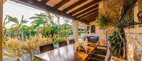 Un patio al aire libre con una mesa de madera y sillas de mimbre, rodeado de exuberante vegetación tropical y un muro de piedra.