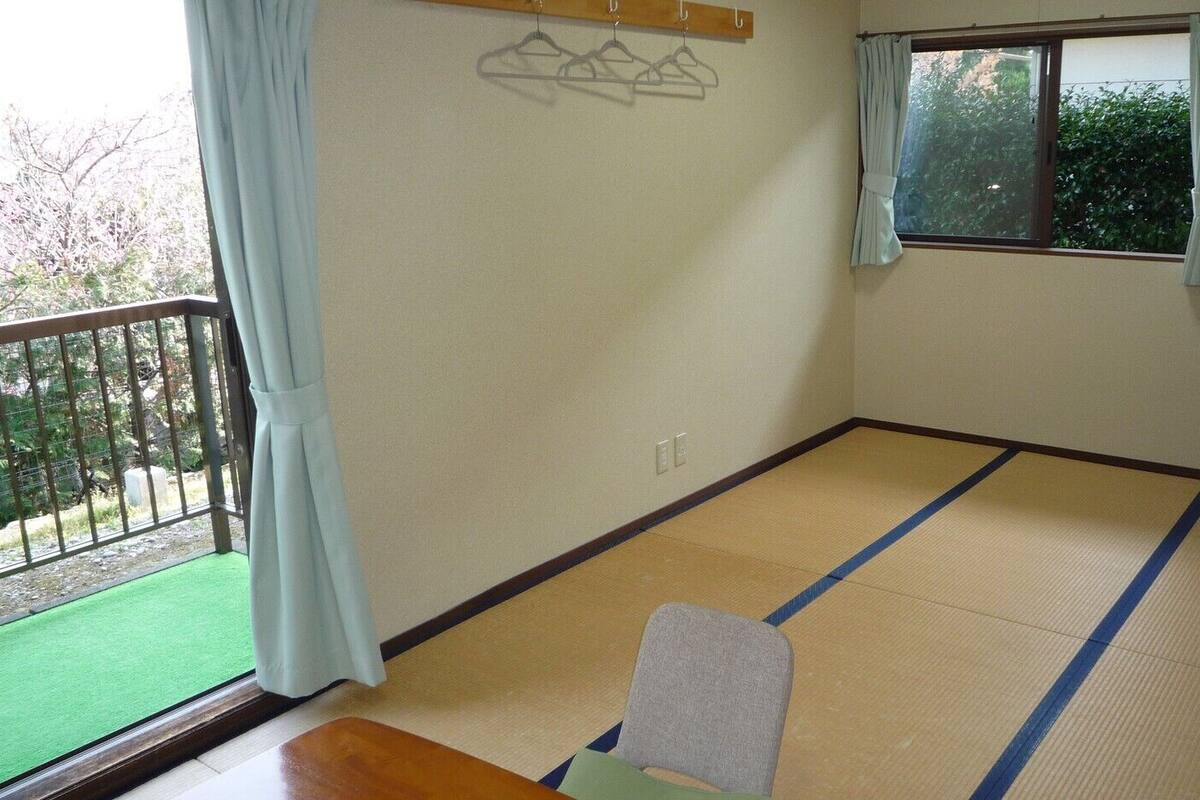 A room with tatami flooring, a wooden table, a chair, a window with curtains, and a view of greenery outside.