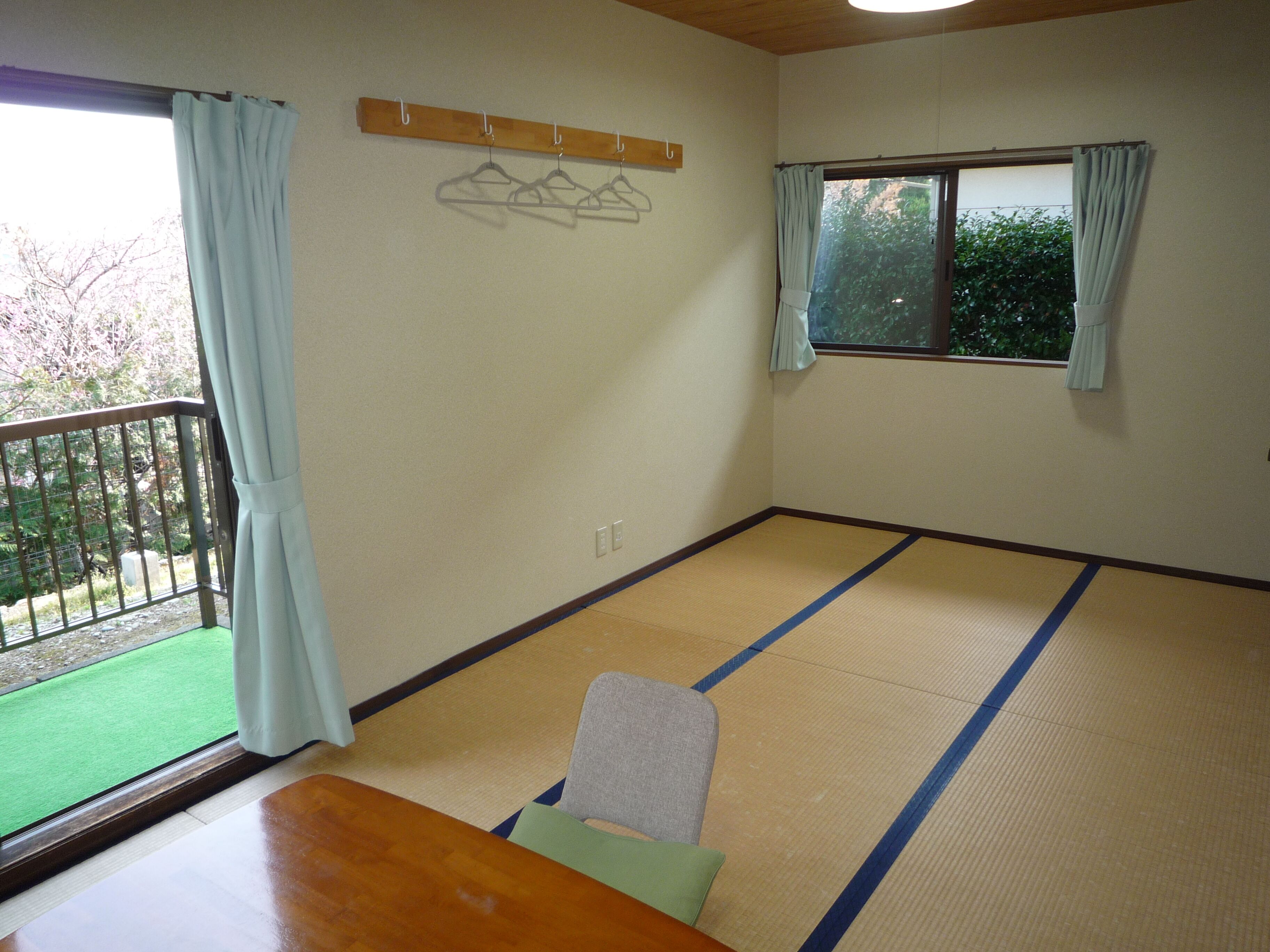 A room with tatami flooring, a wooden table, a chair, a window with curtains, and a balcony with a railing.