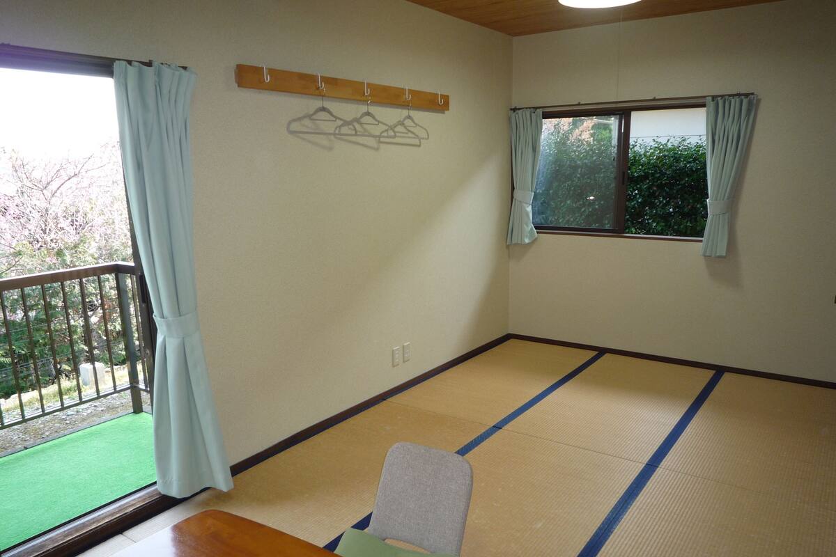 A room with tatami flooring, a wooden table, a chair, a window with curtains, and a balcony with a railing.