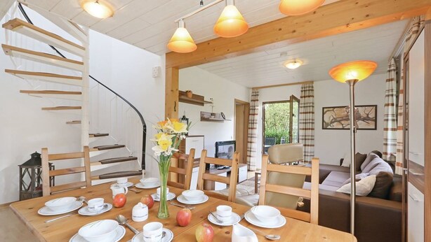 Un comedor con una mesa de madera dispuesta para cuatro personas, rodeada de sillas de madera. Hay un florero con flores sobre la mesa, y manzanas sobre el mantel. Se ve una escalera con escalones de madera a la izquierda.