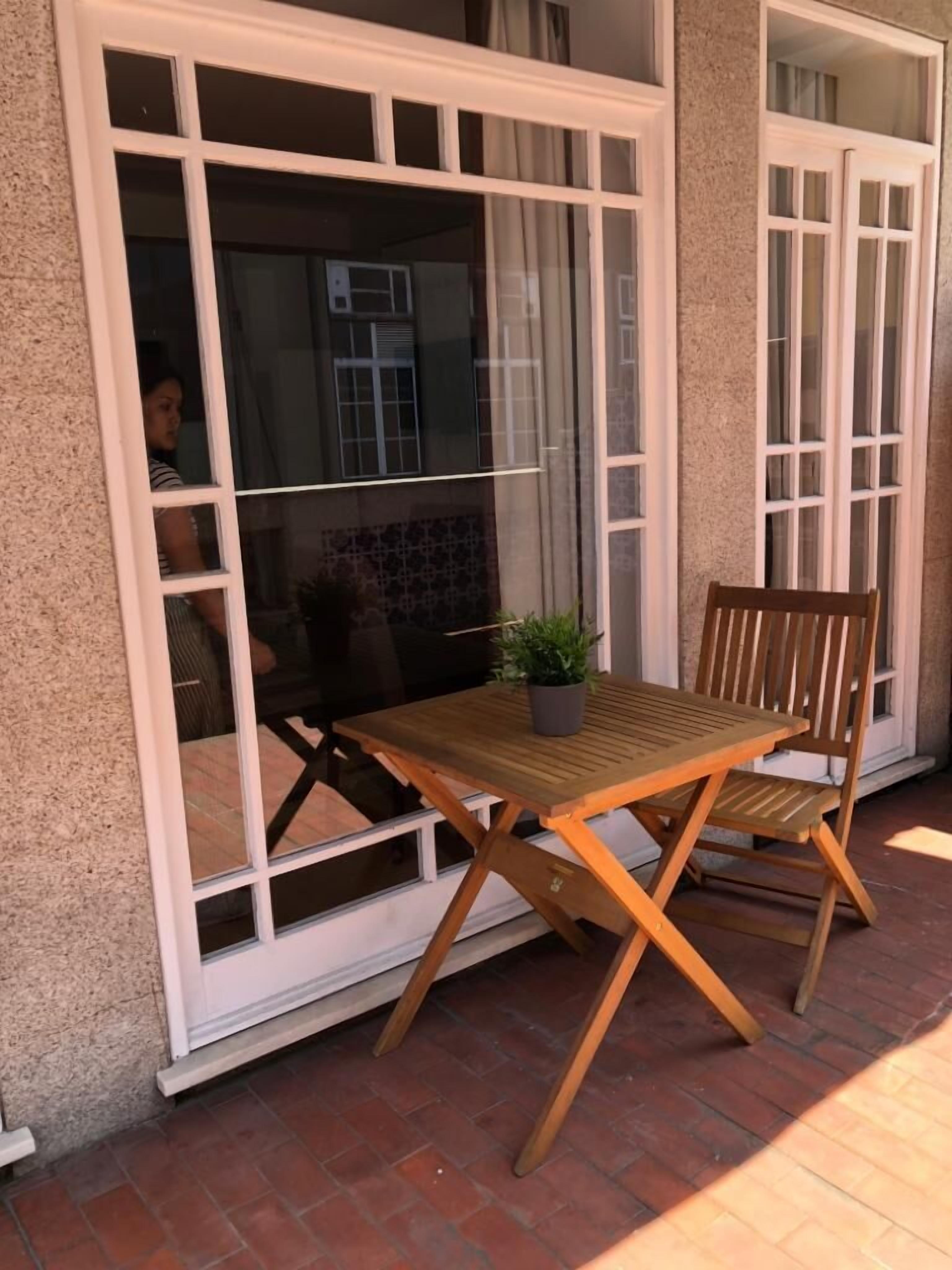 Un patio con una mesa y sillas de madera, una planta en maceta y vistas a través de una puerta de cristal.