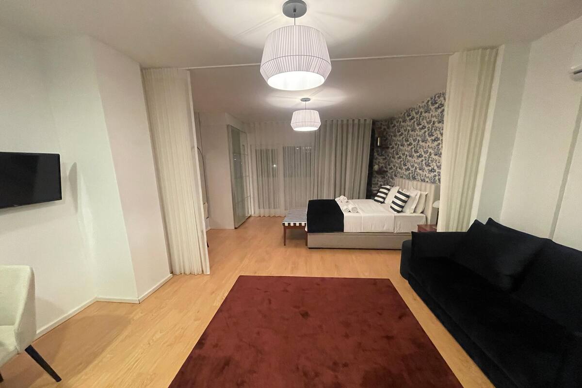 Una habitación de hotel moderna con una cama grande, un sofá negro, un tapete rojo y un televisor montado en la pared.