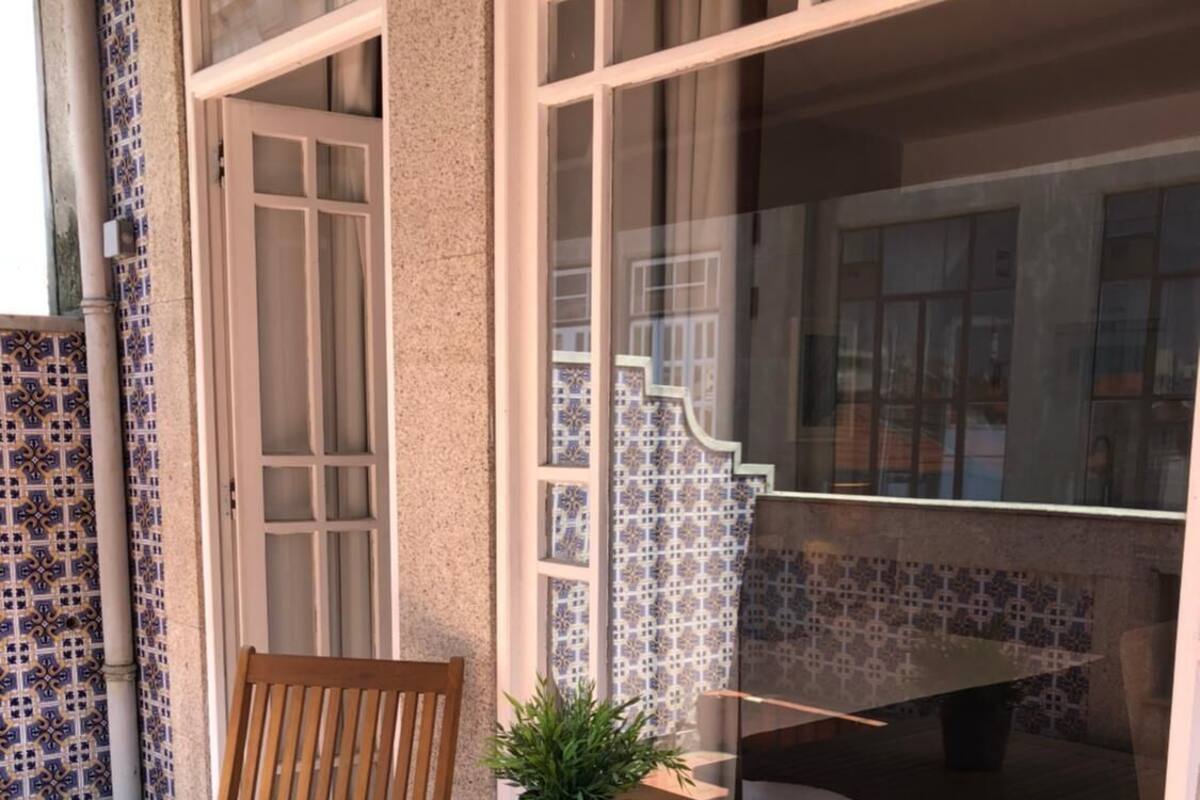 Mesa y sillas de madera al aire libre con una planta en maceta, frente a un edificio con paredes de azulejos estampados y ventanas grandes.
