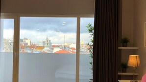 Habitación de hotel con un amplio ventanal corredero, una cama, una mesita de noche con lámpara y vistas a la ciudad.