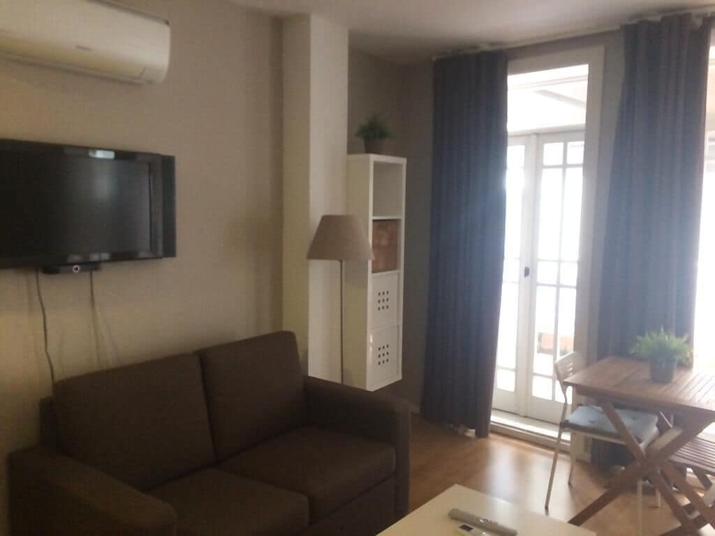A living room with a sofa, a TV mounted on the wall, a table with chairs, and a sliding glass door leading to an outdoor area.