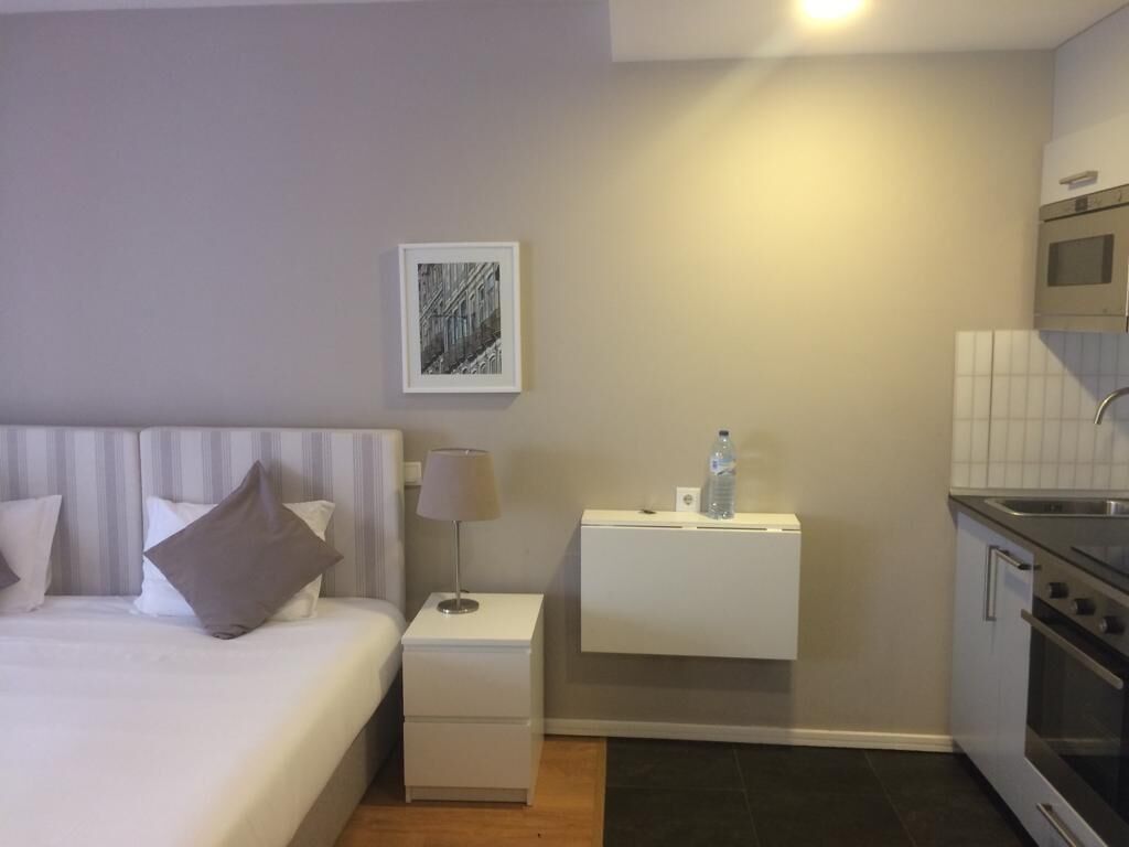 Une chambre d’hôtel comprenant un lit, une table de chevet, une lampe, une bouteille d’eau et un coin cuisine avec une cuisinière et un four.