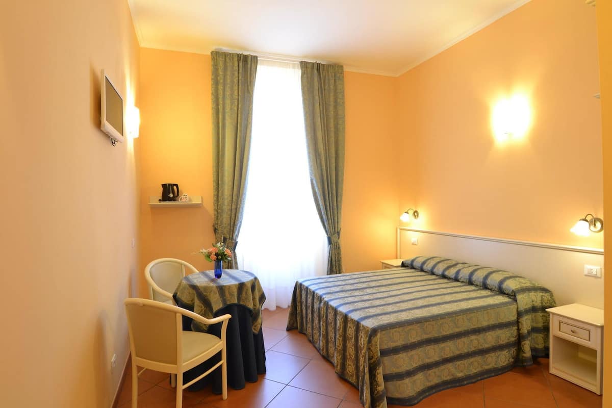 Quarto de hotel pequeno com uma cama de solteiro, mesa de cabeceira, uma cadeira, uma mesinha com abajur e uma janela com cortinas.
