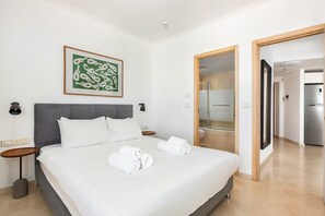 Une chambre d’hôtel avec un grand lit, une table de chevet, une œuvre d’art accrochée au mur et une salle de bain visible à travers une porte ouverte.