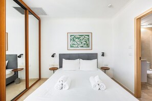 Ein Hotelzimmer mit einem Bett, zwei Nachttischen, einem Spiegel, einem gerahmten Kunstwerk an der Wand und einem durchgehenden Türspalt sichtbaren Badezimmer.