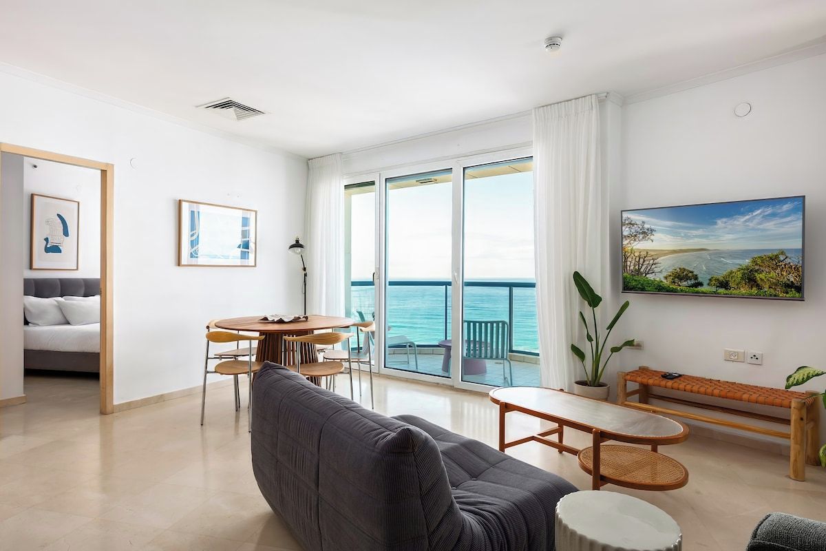 Un salon moderne avec un canapé, une table à manger et des chaises, un balcon offrant une vue sur la mer et un tableau accroché au mur.