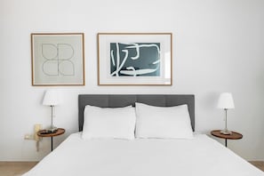 Ein ordentlich bezogenes Bett mit weißen Leinen, zwei Nachttische mit Lampen und zwei gerahmte abstrakte Kunstwerke an der Wand.