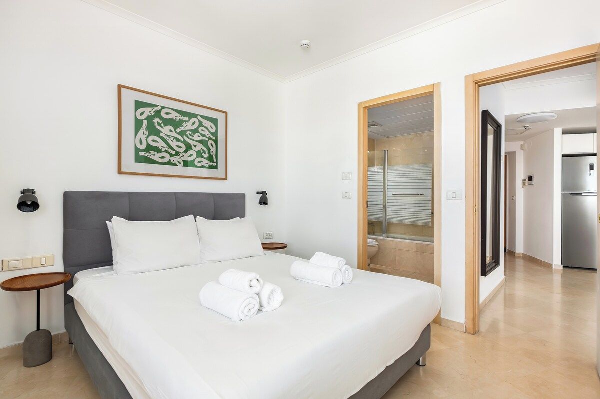 A hotel room with a large bed, a bedside table, a wall-mounted artwork, and a bathroom visible through an open door.