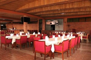A spacious dining area with multiple tables and chairs, a brick wall, and a wooden ceiling. - Sainte Famille Hotel (Kigali)