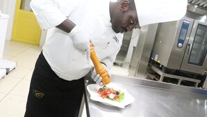 Un chef con uniforme blanco y delantal negro preparando alimentos en una cocina de acero inoxidable.