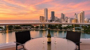 Mesa redonda con dos sillas, una botella de vino y dos copas en un balcón con vistas a un horizonte urbano al atardecer.