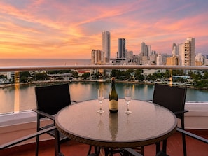 Mesa redonda con dos sillas, una botella de vino y dos copas en un balcón con vistas a un horizonte urbano al atardecer.