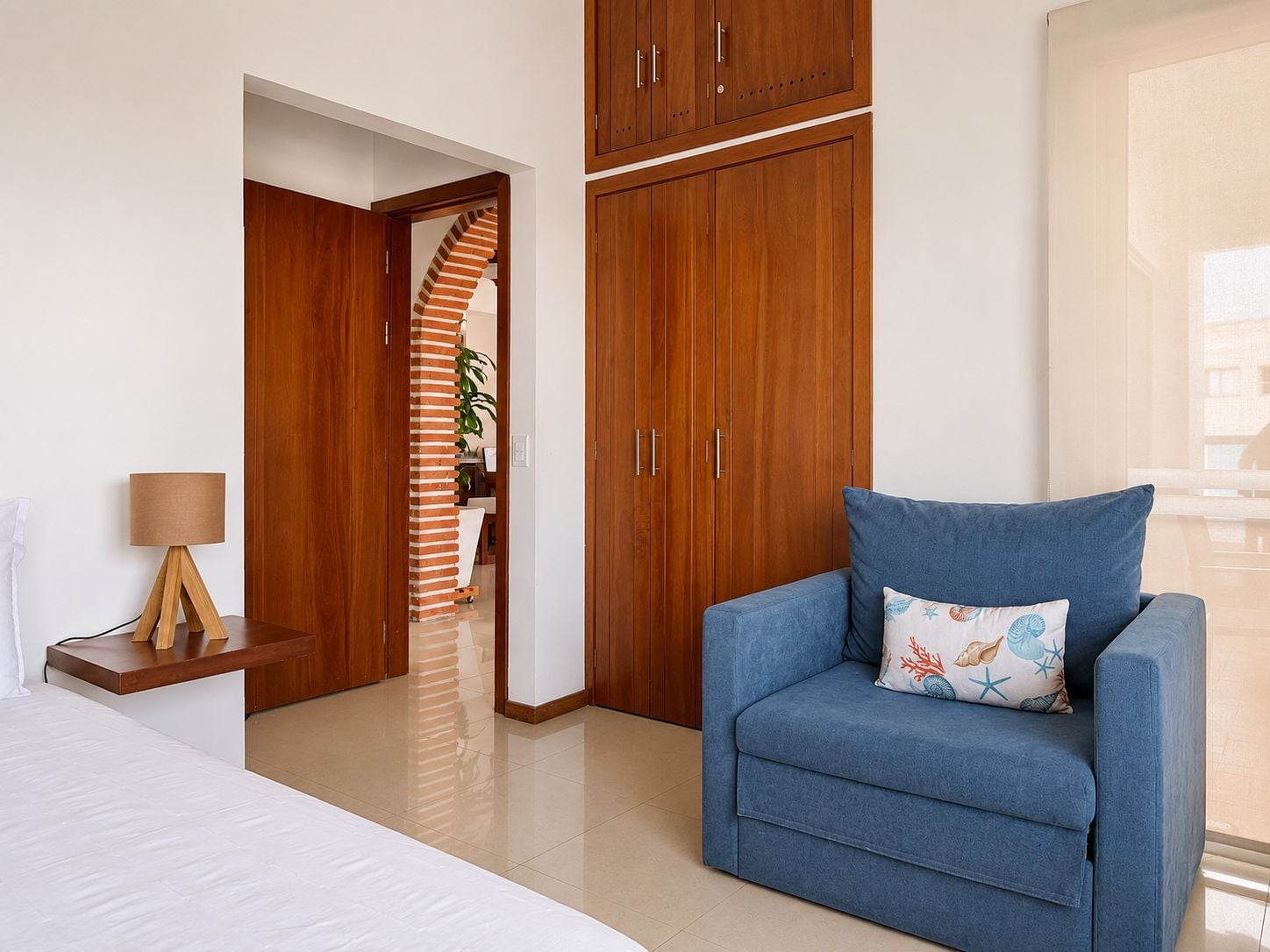 A hotel room with a bed, a blue armchair, wooden wardrobes, a lamp, and a view of the outside.