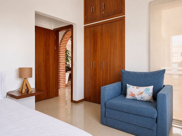 A hotel room with a bed, a blue armchair, wooden wardrobes, a lamp, and a view of the outside.