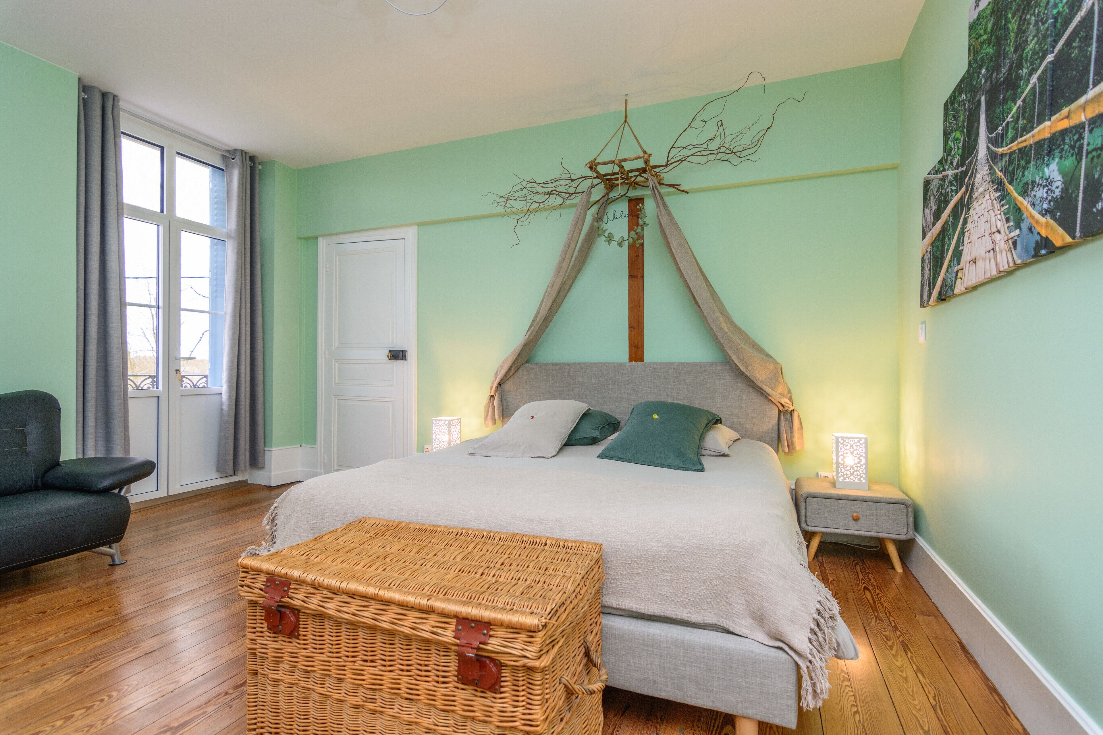 Un dormitorio con cama, mesita de noche, baúl de mimbre, una silla y ventana con cortinas.