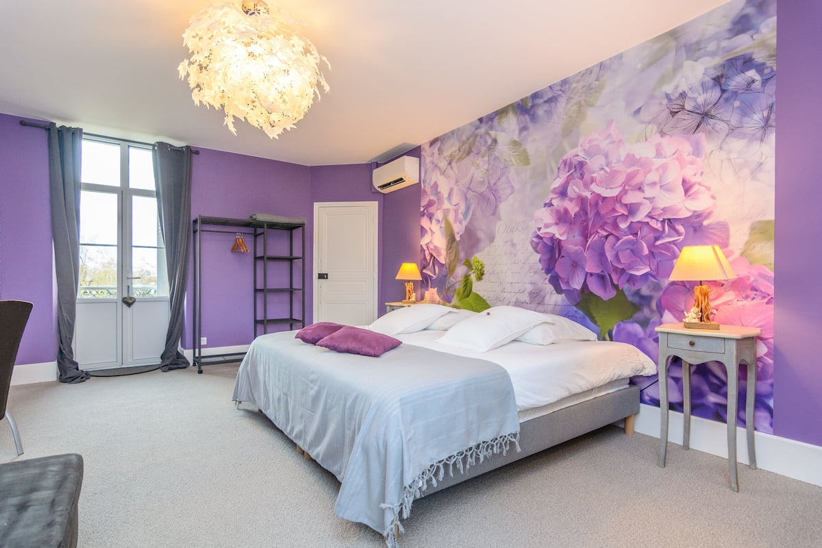 A bedroom with a bed, two bedside lamps, a chandelier, a window with curtains, and a floral wall mural.