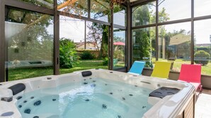 Een bubbelbad met glazen omheining, omringd door een terras met kleurrijke stoelen en uitzicht op een tuin en huizen.