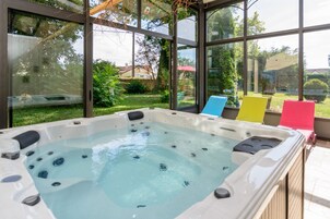 Un jacuzzi con cerramiento de vidrio, rodeado por una terraza con sillas coloridas y vistas a un jardín y casas.