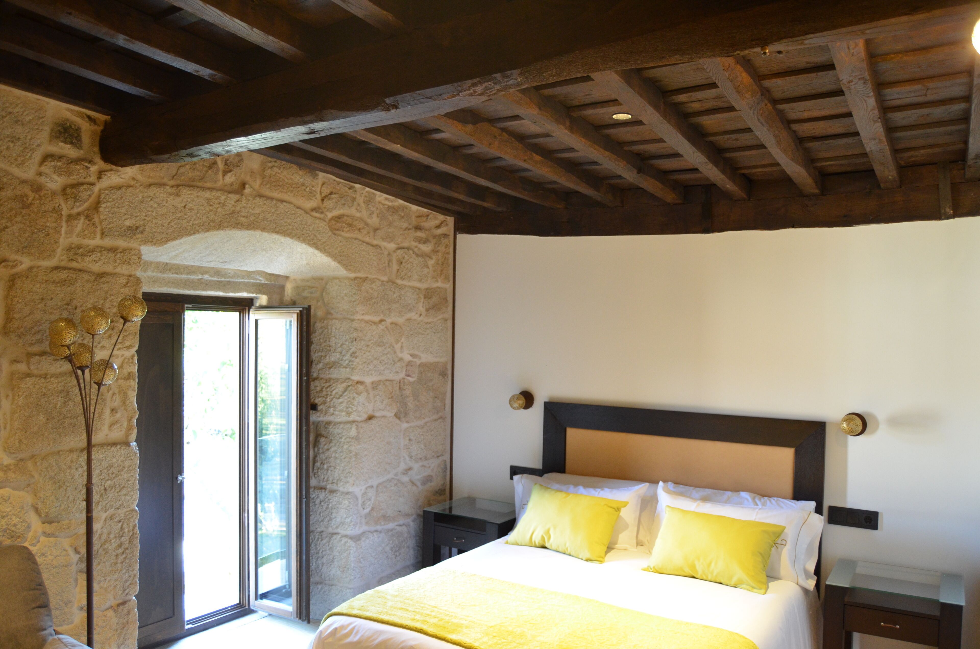 Un dormitorio con pared de piedra, techo de madera, cama con ropa de cama amarilla y una mesita de noche.