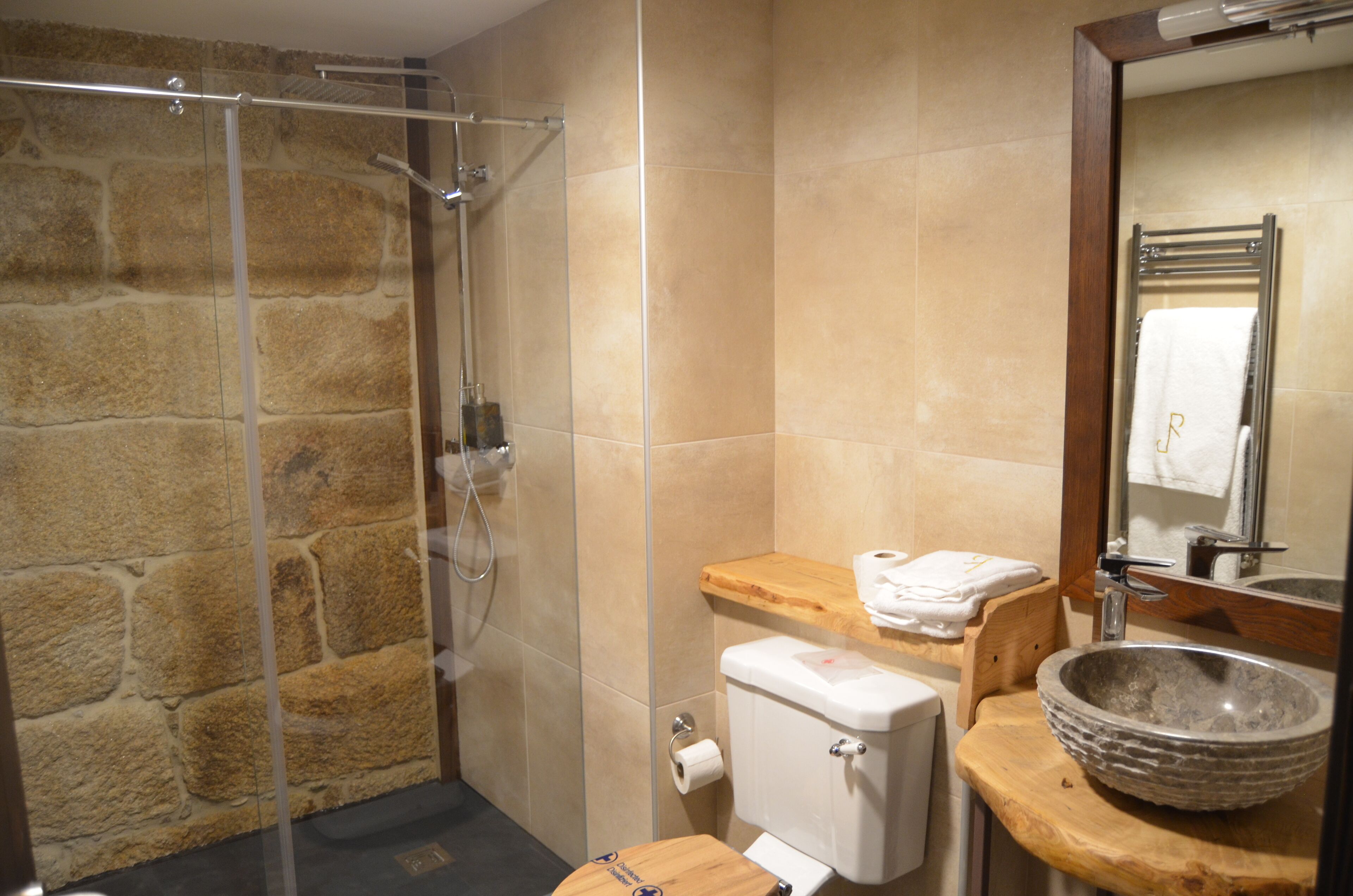 A bathroom with a stone wall, a glass shower enclosure, a wooden vanity, a sink, and a toilet.