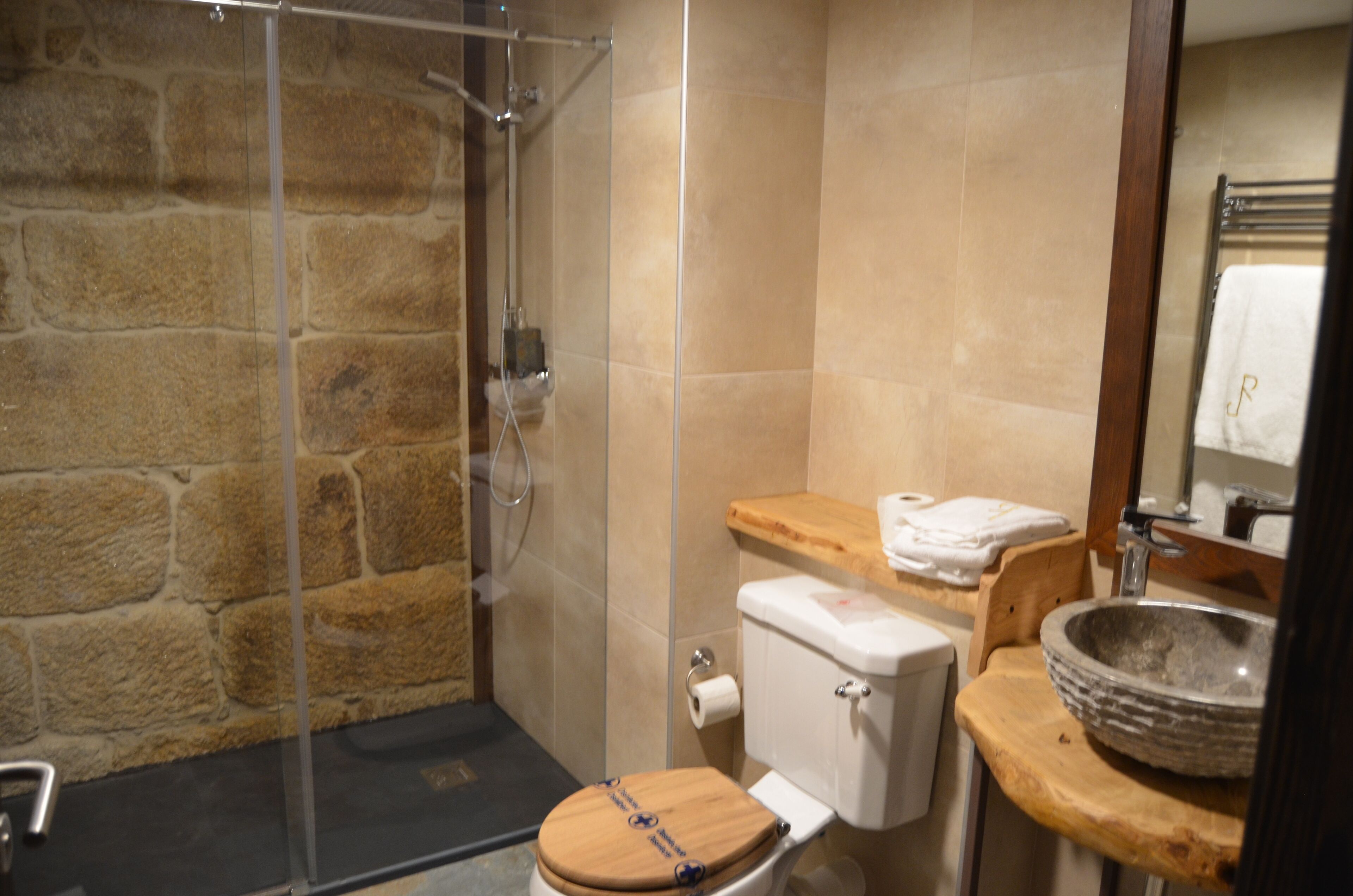 Un baño con pared de piedra, inodoro de madera, lavabo con recipiente de piedra y cabina de ducha con mampara de vidrio.