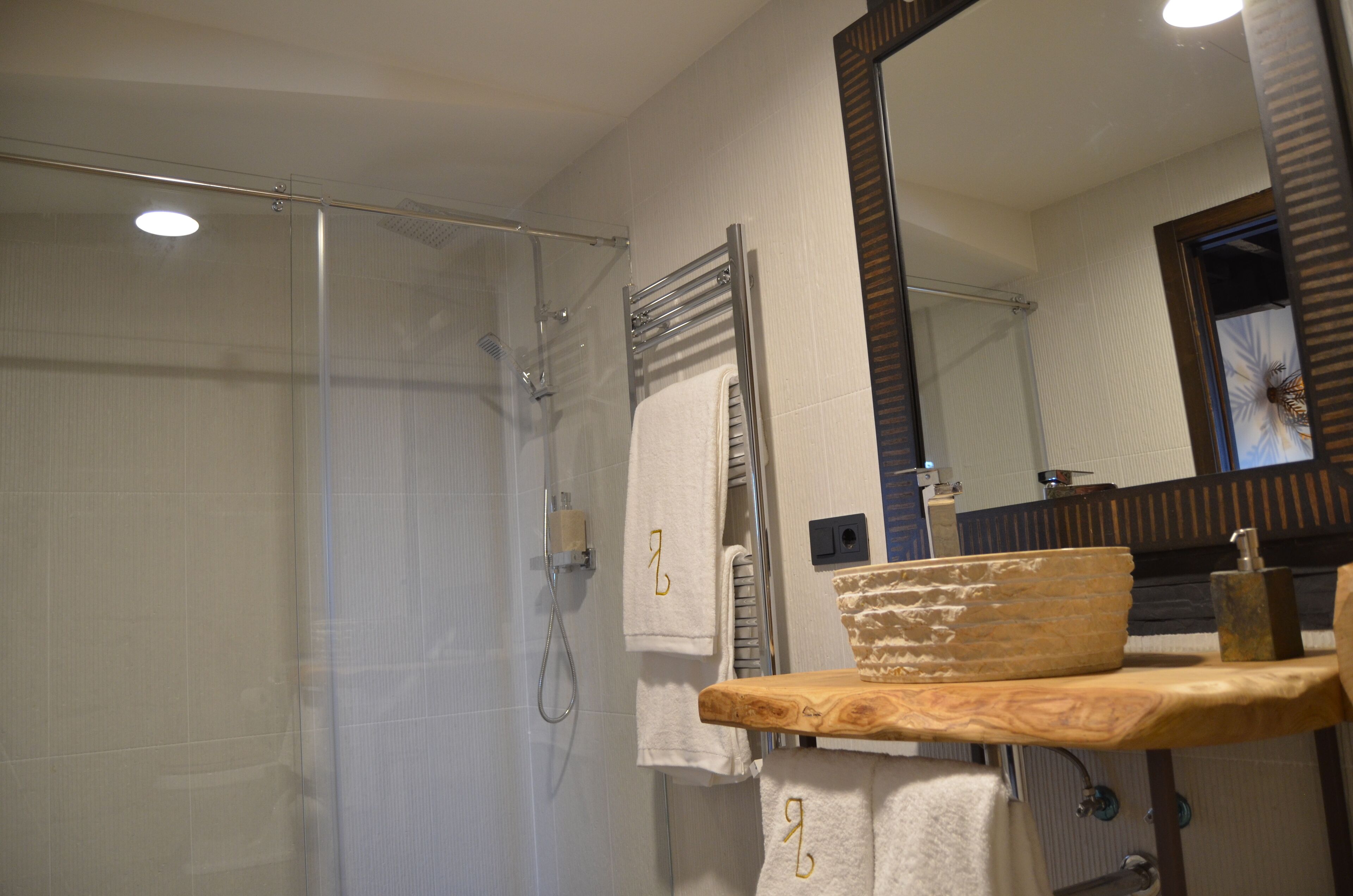A bathroom with a glass shower enclosure, a wooden vanity with a soap dispenser, and a towel rack with towels.