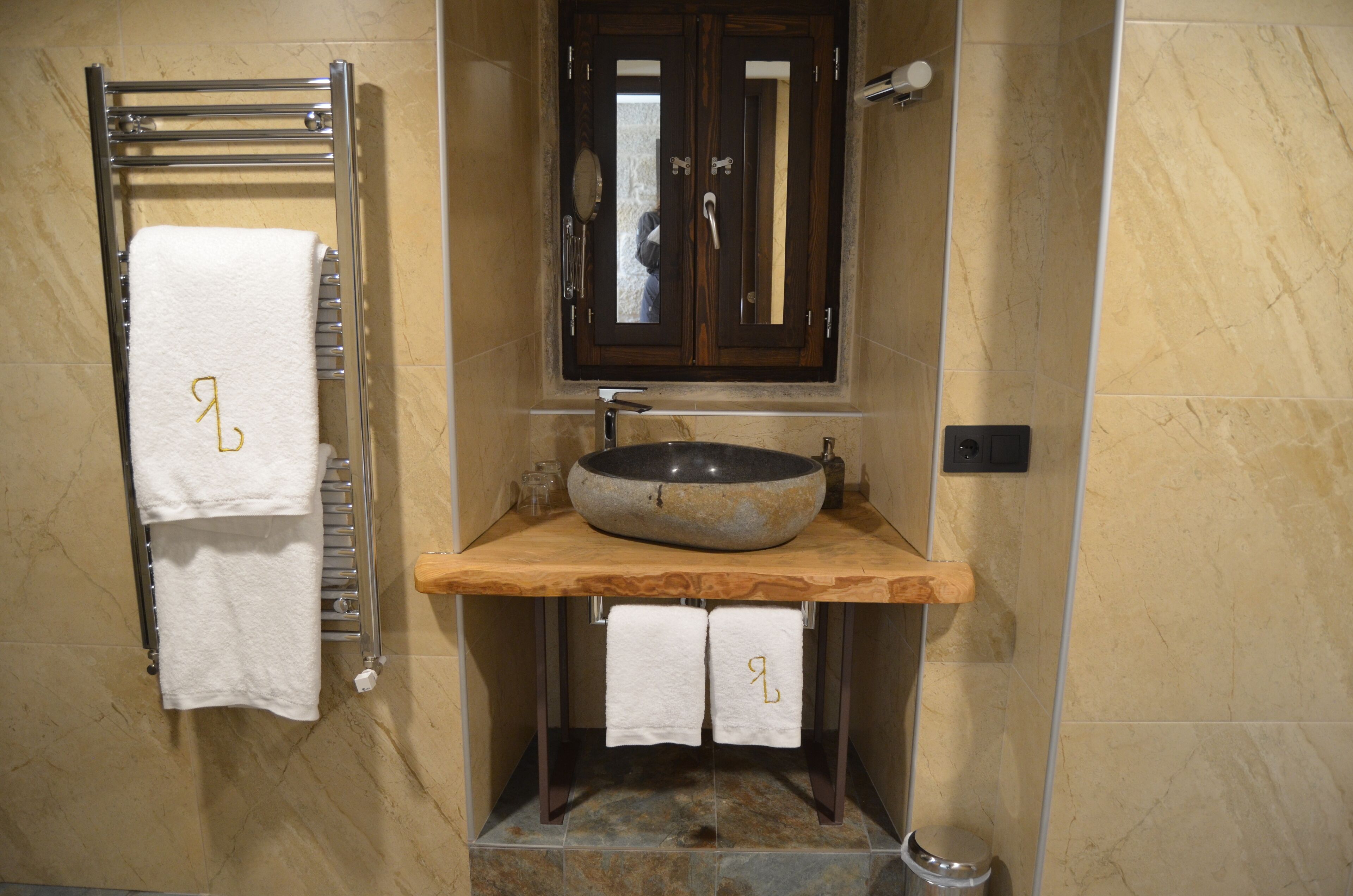 Un bany amb una paret revestida de marbre, un tocador de fusta amb un gran lavabo, un penjallina de tovalloles i un mirall sobre el lavabo.