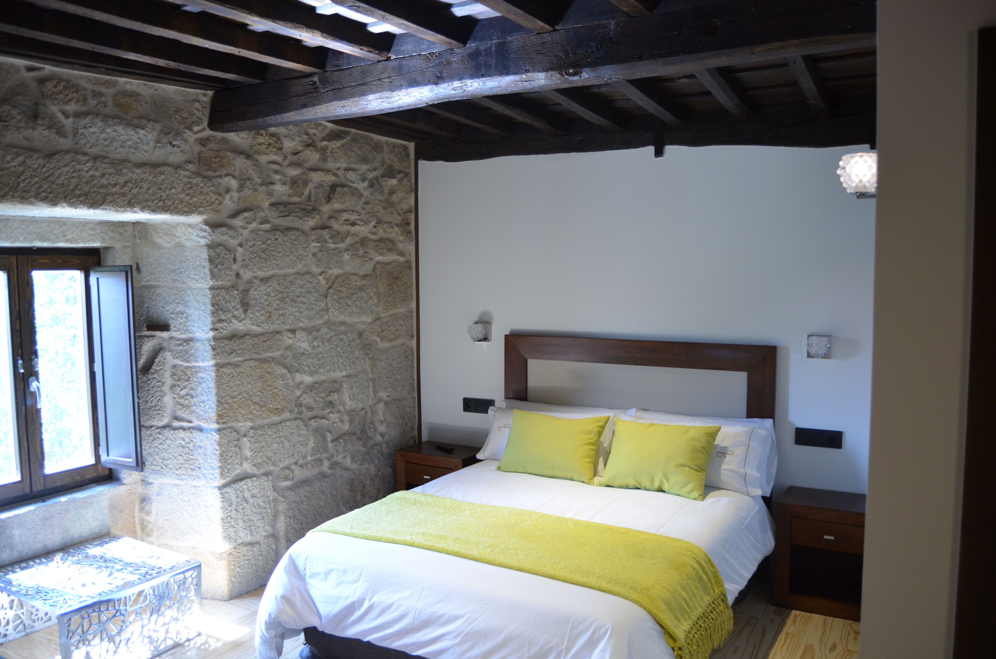 A bedroom with a stone wall, a wooden ceiling, a bed with white and yellow bedding, and a nightstand.